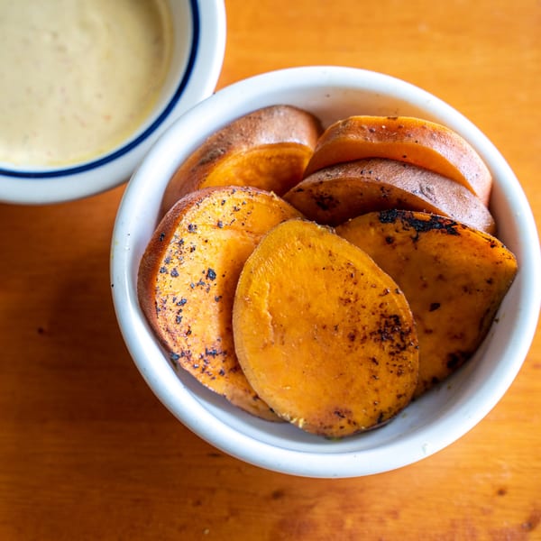 A white bowl filled with sliced, roasted sweet potatoes, next to a bowl of creamy sauce.