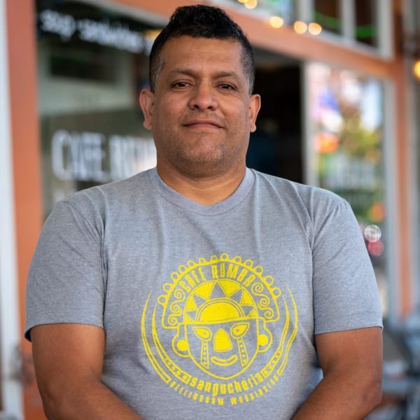 Man in a gray Cafe Rumba t-shirt with a yellow Peruvian design, standing outside.