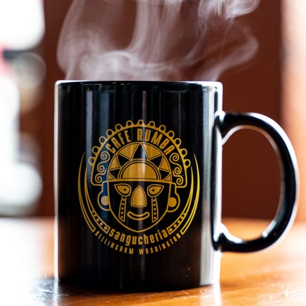 Steaming black Cafe Rumba mug with a gold Peruvian-inspired logo on a wooden surface.