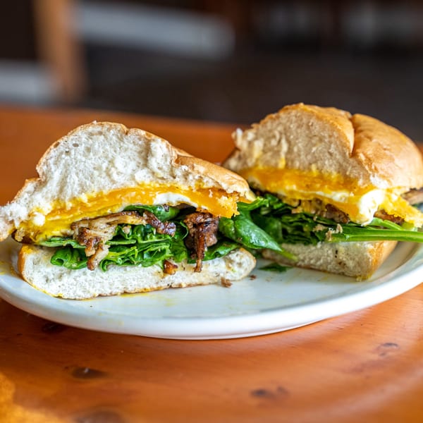 A Peruvian sandwich, cut in half, with fried egg, greens, and caramelized onions on a white plate.