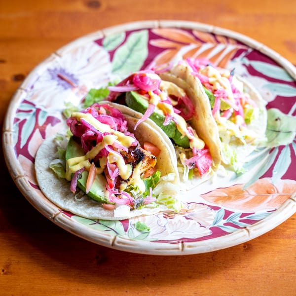 Three Peruvian tacos with grilled fish, avocado, pickled red onions, and yellow sauce on a floral plate.